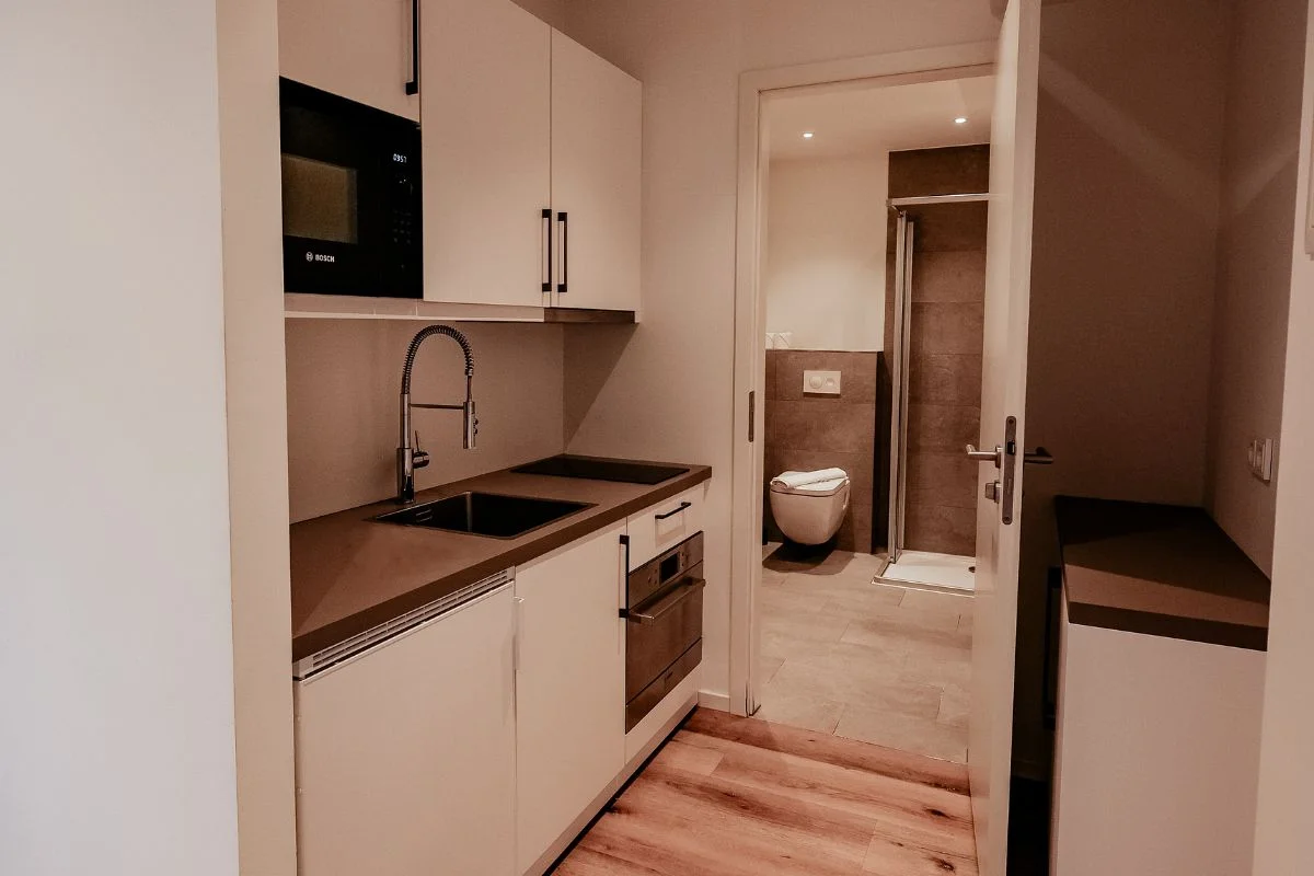 Kompakte Küchenzeile mit Spüle, Herd und Mikrowelle, die an ein Badezimmer grenzt. Durch die offene Tür sieht man die Dusche und die Toilette.