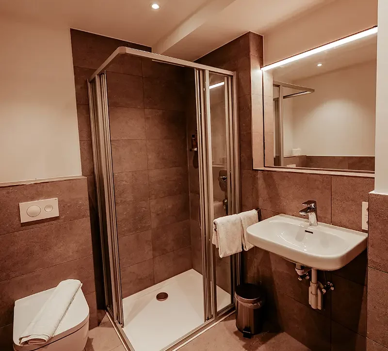 Ein modernes Badezimmer mit großen grauen Fliesen, einer Toilette, Waschbecken und einer Dusche sowie einem großen beleuchteten Spiegel.