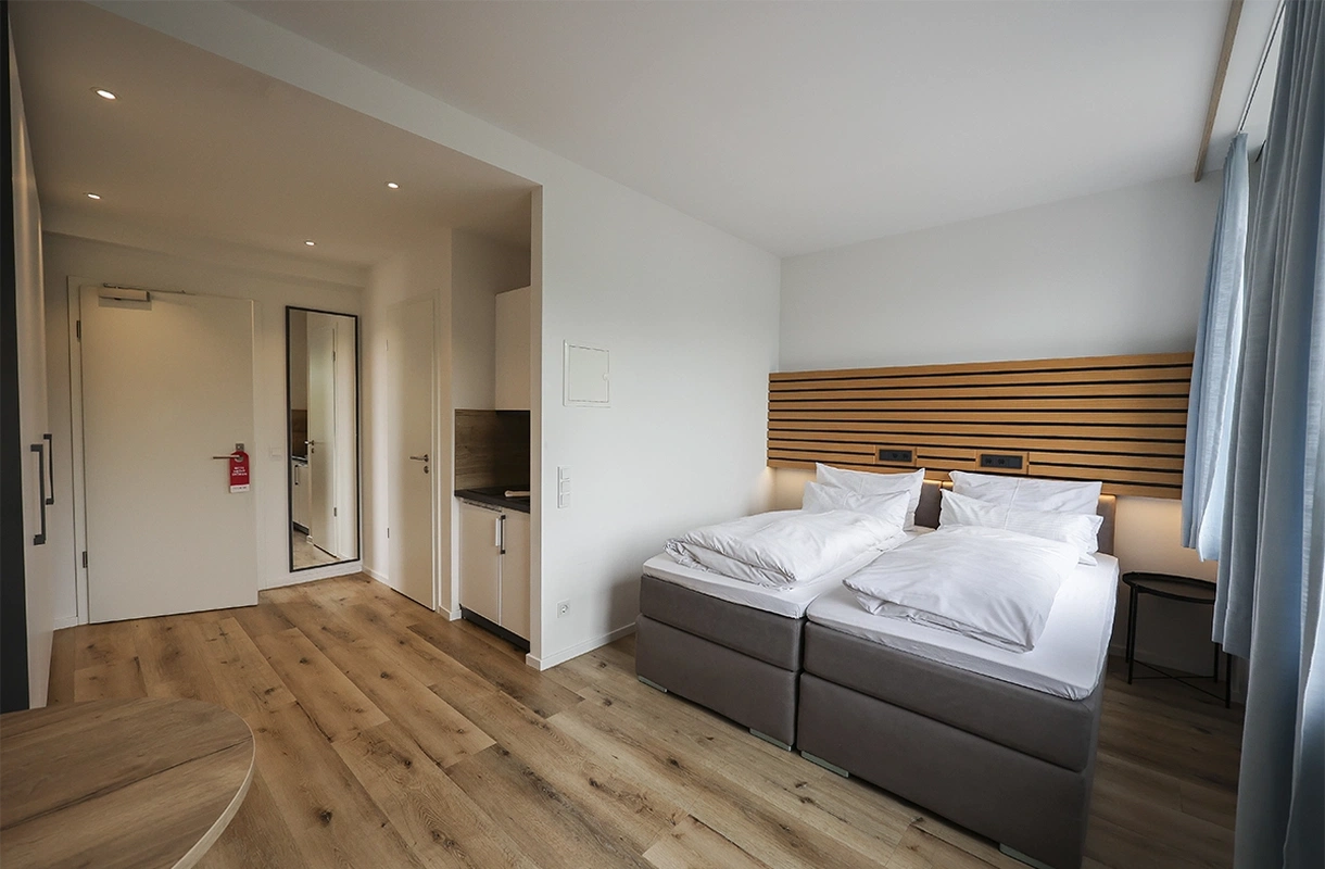 Ein helles und modernes Double Apartment mit zwei Betten, einer kleinen Küchenzeile, einem großen Spiegel und Holzpaneelen an der Wand.