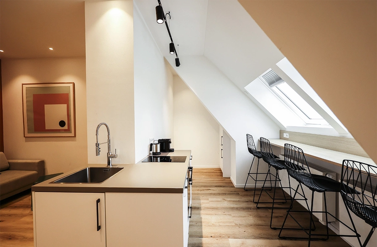 Eine moderne, aufgeräumte Küche mit Barhockern an einer Theke unter einem Dachfenster und einem Kunstwerk an der Wand bei Zählwerk Apartments.